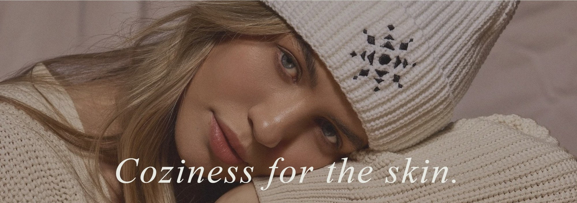 Coziness for the skin. A woman in a cream knit beanie and sweater rests her head on her arm.