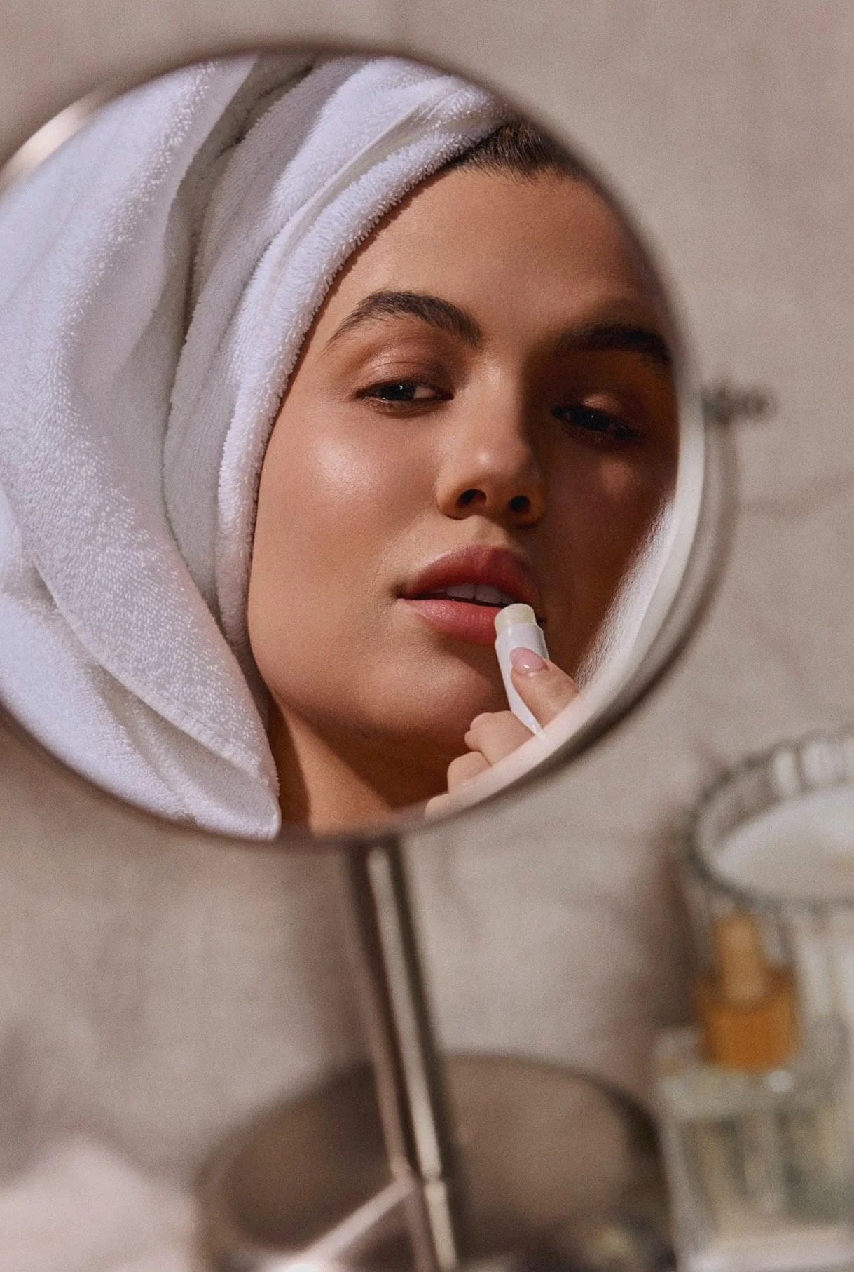 Woman with a towel wrapped around her hair applying Lun lip balm, seen in a round mirror